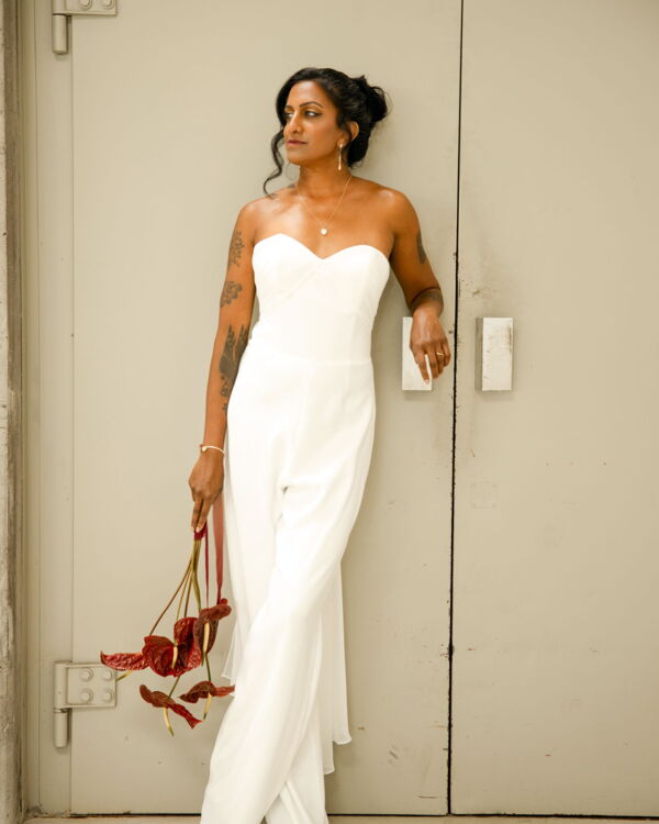 Ganzkörperporträt der Braut im weißen Outfit, lässig an einer Tür lehnend mit roten Anthurien